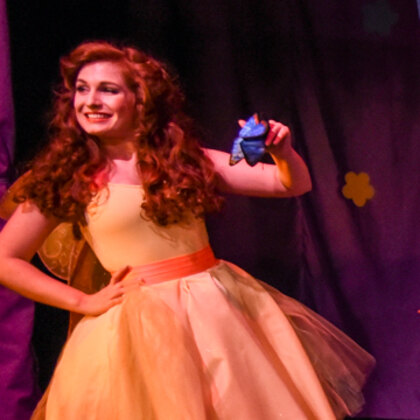 Lulu in a gold dress, smiling, holding a small blue creature on the stage.