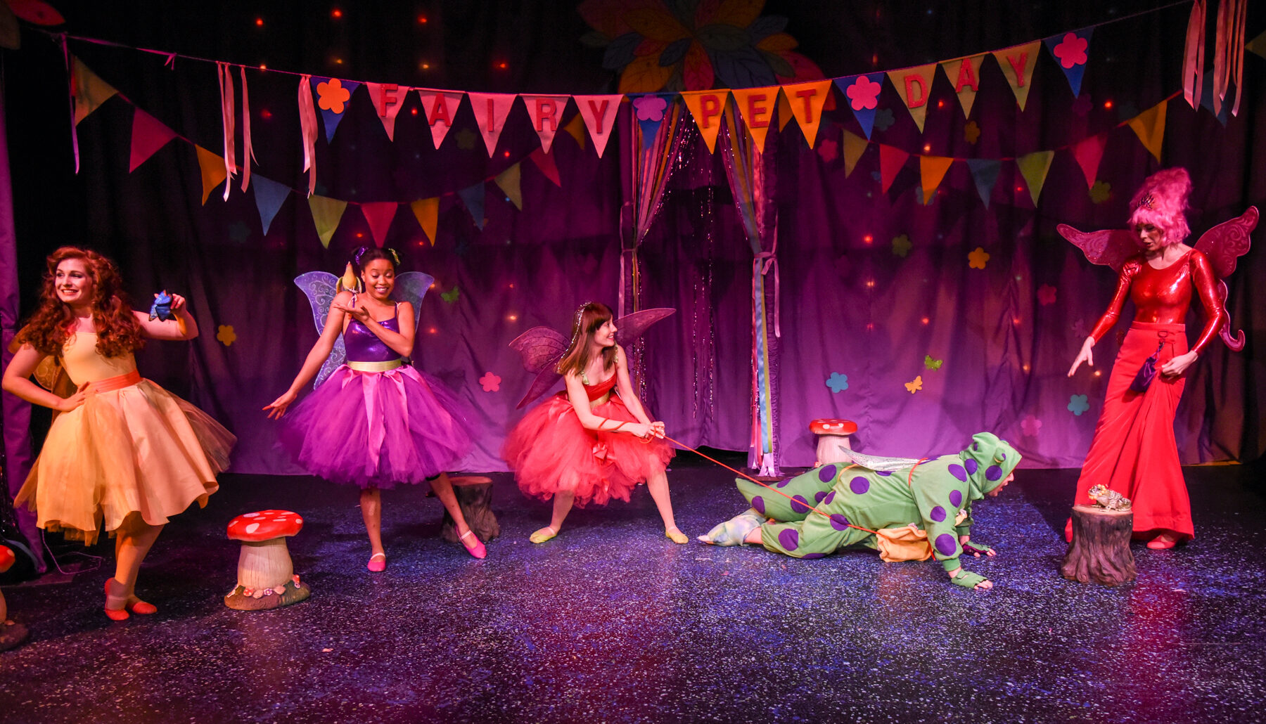 Fairy Pet Day on stage: bunting reading FAIRY PET DAY, three fairies watching as Twinkle hauls on Scruffy's leash and the Fairy Godmother looks on.