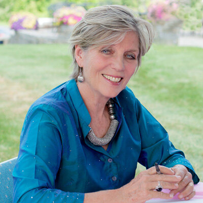Katharine Holabird, author, smiling at a garden table with a pen in hand.